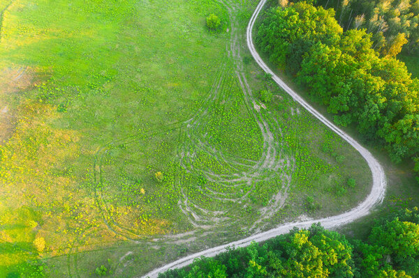 Ground highway dirt road on the lawn near the trees on the edge of the green forest in the summer at sunset with long shadows - aerial shot with a place for text