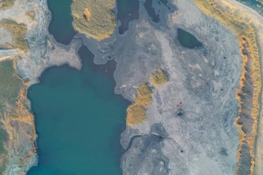 Bir madencilik göletinin havadan soyutlanması. Gri, çatlak atık, derin mavi-yeşil su ve sarı sazlıklar ile çelişiyor. Bu durum ciddi endüstriyel kirlilik ve çevresel hasarı gösteriyor..