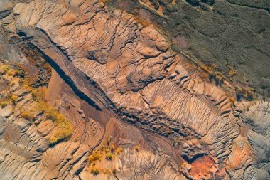 Heykeltraş edilmiş, aşınmış endüstriyel atıkların soyut hava görüntüsü. Terracotta kırmızı toprağı yoğun jeolojik akış örüntülerini gösterir, koyu yeşil doğal sınırla keskin bir zıtlık gösterir..