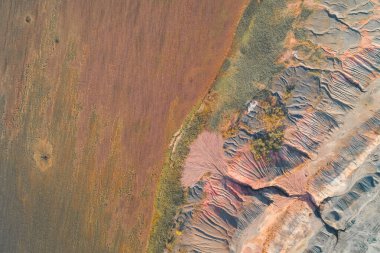 Kahverengi bir tarla ile renkli endüstriyel çorak arazi arasındaki bölünmenin soyut hava aracı görüntüsü. Erimiş kırmızı ve gri kil yamaçlar çevresel hasarı ve şiddetli toprak erozyonunu vurgular..