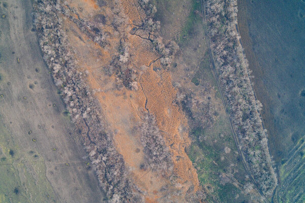 Aerial drone shot of a vibrant red industrial slag pile with cracked erosion patterns. Top down view of mining waste pollution framed by dry shelterbelts. Environmental disaster texture and ferric oxide soil.