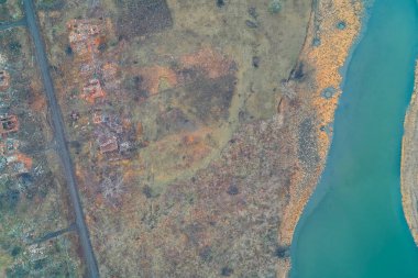 Nehir kıyısındaki yıkık evlerin yukarıdan aşağı görüntüsü. Akan su ve harabe evler arasındaki zıtlık, savaşın nehir kenarındaki toplumlar üzerindeki şiddetli etkisini vurguluyor..