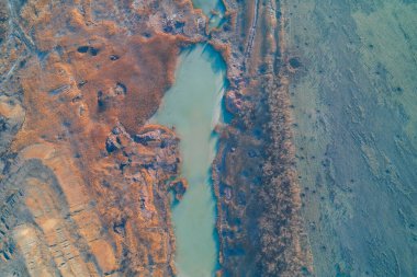 Turkuaz zehirli su havuzunun turuncu kil toprağı ve kurumuş sazlıkları keserken gerçeküstü görüntüsü. Kimyasal kirliliği, taş ocağı kazma yaralarını ve çevresel hasar dokusunu ortaya çıkaran bir distopyan sanayi bölgesi.