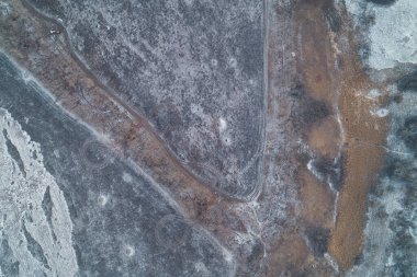 Dolambaçlı toprak bir yol, donmuş toprakta dairesel ağır araç izleri ile işaretlenmiş savunma ormanı şeridini kuşatır. Bu hava kışı manzarası askeri lojistik, devriye rotaları ve taktiksel manevraların stratejik gerilimini yakalar..