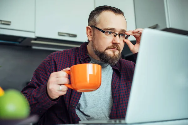 Stylish intelligent man with a beard in modern minimalist kitchen interior. Businessman in a home shirt having breakfast and studying the news looking at laptop.