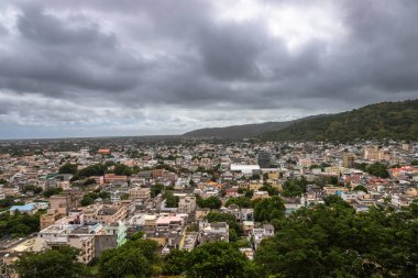 Mauritius 'un başkenti Port Louis' in panoramik manzarası. Arka planda karamsar gökyüzünün altında dramatik bir dağ sırası var. Bu şehir manzarası, modern mimari ve doğal manzara arasındaki karşıtlığı vurgular, başyazı ve turlar için idealdir.