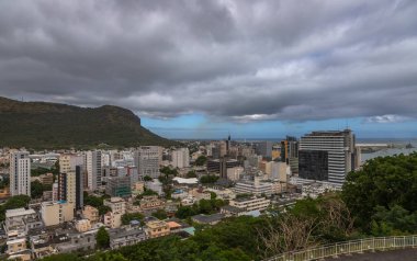 Mauritius 'un başkenti Port Louis' in panoramik manzarası. Arka planda karamsar gökyüzünün altında dramatik bir dağ sırası var. Bu şehir manzarası, modern mimari ve doğal manzara arasındaki karşıtlığı vurgular, başyazı ve turlar için idealdir.