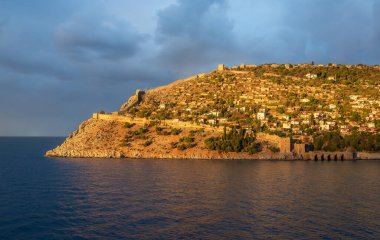 Altın gün batımı ışığı Alanya Şatosu 'nu ve Alanya Türkiye' deki Akdeniz 'in üzerindeki antik kale duvarlarını aydınlatarak tarihi mimari ve manzaralı kıyı güzelliğini gözler önüne seriyor.