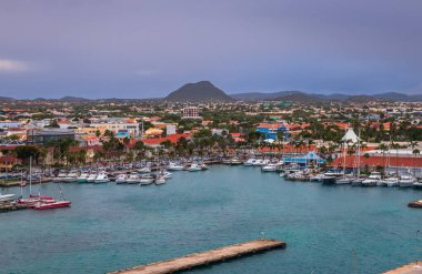 Oranjestad, Aruba 'nın panoramik manzarası, renkli Hollanda Karayipleri mimarisi, palmiye ağaçları ve dramatik akşam gökyüzü altında turkuaz deniz manzarası