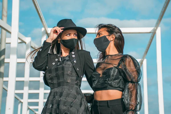 İki genç kızın birlikte poz verdiği moda fotoğrafçılığı. Biri açık tenli, sarışın, diğeri kahverengi tenli, siyah saçlı ve ikisi de siyah koruyucu maskeli..