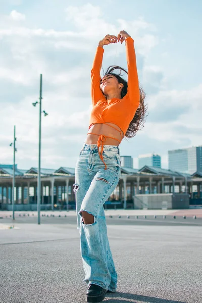 Cılız, koyu tenli genç bir kadınla moda fotoğrafçılığı. Turuncu bir bluz ve kot pantolon giymişti, gündüz vakti sokakta..