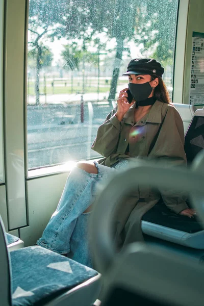 A young woman with brown skin wearing a black protective face mask sits inside public transport using her mobile phone.