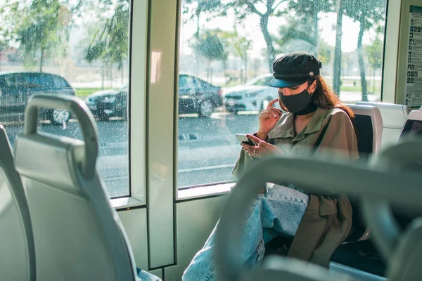 A young woman with brown skin wearing a black protective face mask sits inside public transport using her mobile phone.