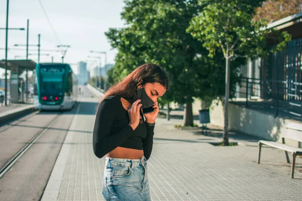 A young woman with dark skin in a black protective face mask waiting near the tram lines using her mobile phone.