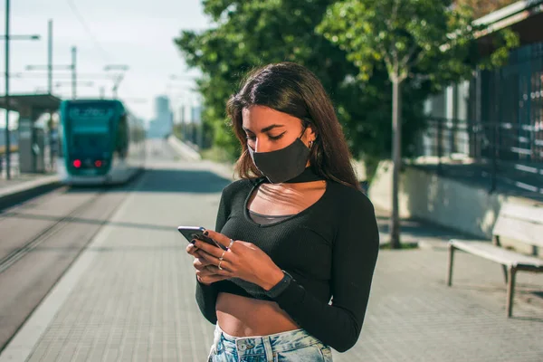 A young woman with dark skin in a black protective face mask waiting near the tram lines using her mobile phone.