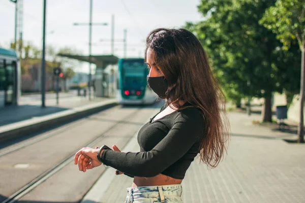 Siyah koruyucu maskeli, kahverengi tenli genç bir kadın güpegündüz tramvay hattının yanında dijital saatine bakıyor..
