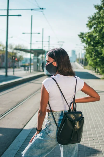 Siyah koruyucu maskeli, koyu tenli genç bir kadın güpegündüz tramvay hattının yanında sırtı yana dönük bekliyor..