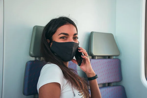 Kahverengi tenli, siyah koruyucu maskeli genç bir kadın toplu taşıma araçlarında cep telefonunu kullanıyor..