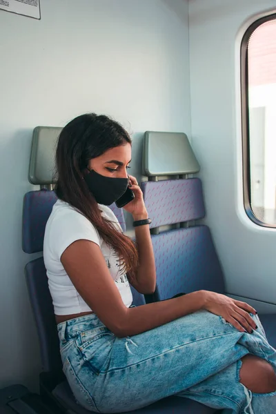 A young woman with brown skin wearing a black protective face mask using the mobile phone in public transport.