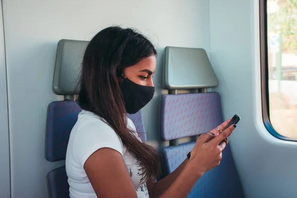 A young woman with brown skin wearing a black protective face mask using the mobile phone in public transport.