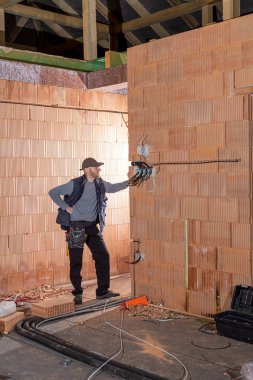 İşteki bir elektrikçi, kil tuğlalardan yapılmamış eve prizler yerleştirmeye hazır. İşçi inşaat halindeki evin içindeki kablolara hazır..