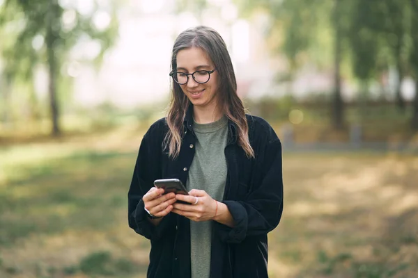 Mladá žena s brýlemi čtení zprávu na telefonu, zatímco stojí v parku — Stock fotografie