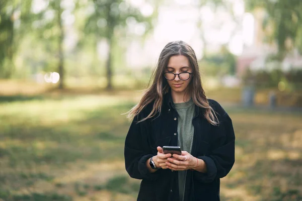 Mladá žena s brýlemi čtení zprávu na telefonu, zatímco stojí v parku — Stock fotografie