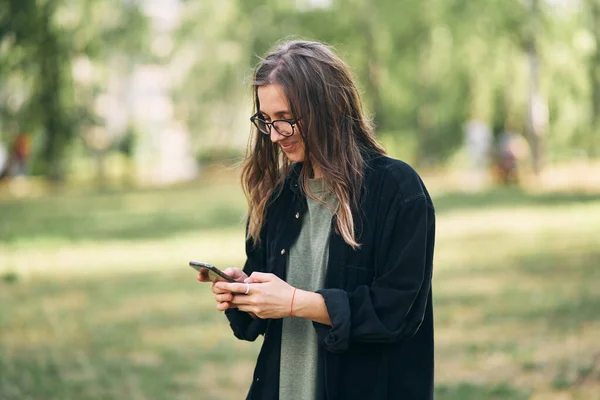 Jeune femme avec des lunettes lisant un message sur son téléphone tout en se tenant dans le parc — Photo