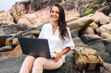 Genç, başarılı, gülümseyen bir kız dizüstü bilgisayar kullanıyor, kayalık bir deniz kıyısında oturuyor. blogger, freelancer, online çalışma