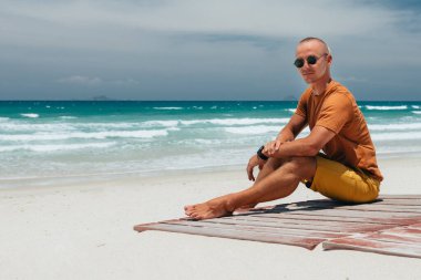 Adam yaz tatilinde tropik bir plajda oturuyor. Kumlu bir sahili olan panoramik bir deniz manzarası, yaz günü, beyaz bulutlu mavi bir gökyüzü. İnanılmaz bir manzara. Yer kopyala