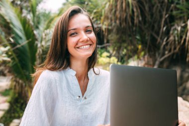 Çok güzel gülümseyen bir kızın yakın plan portresi iş yerinde dizüstü bilgisayarıyla oturuyor. Arka planda tropik bitki örtüsü bulanıklaşır. Serbest kız.