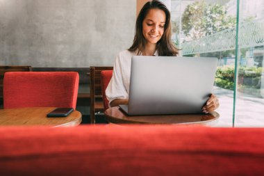 İş kadını ofisinde değil. Kafede otururken dizüstü bilgisayarı için çalışıyor. Uzaktan çalışmada modern teknolojileri kullanmak. Büyük bir pencerenin yanındaki ahşap bir masada oturuyor. Defterde mesaj okuyor. Boşluğu kopyala