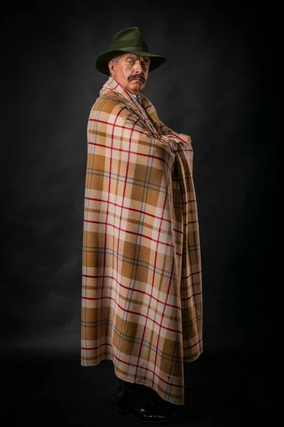 A mature gray-haired man and a gentleman with a mustache, a Latinos in a hat and a light checkered poncho, cape, cloak on a dark background, who looks with an attentive and piercing gaze