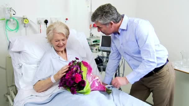 Cuidado hijo trayendo flores sénior mamá hospital cama — Vídeo de stock