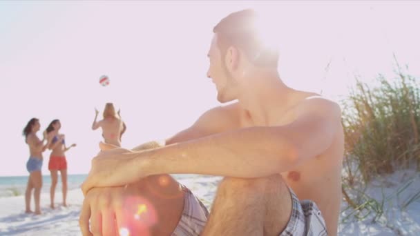 Adolescente sentado en la playa mientras sus amigos juegan pelota — Vídeos de Stock
