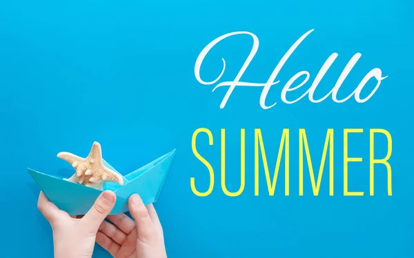 Bonjour l'été. Carte d'été avec un bateau en papier et une étoile de mer sur fond bleu dans les mains d'un enfant. Voyage à la mer et vacances en été — Photo