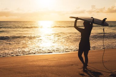 Gün batımı plajında güneş ışığı arka planında sörf tahtalarını taşıyan sörfçü kadın silueti.