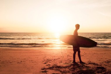 Gün batımı plajında güneş ışığı arka planında sörf tahtalarını taşıyan sörfçü silueti.