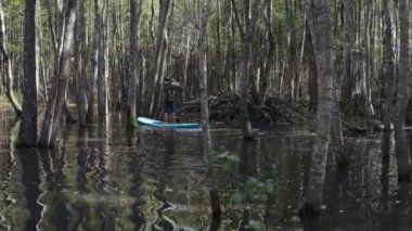 Küçük bir göldeki ağaçların arasında, tahtanın üzerinde yüzen genç bir adam.