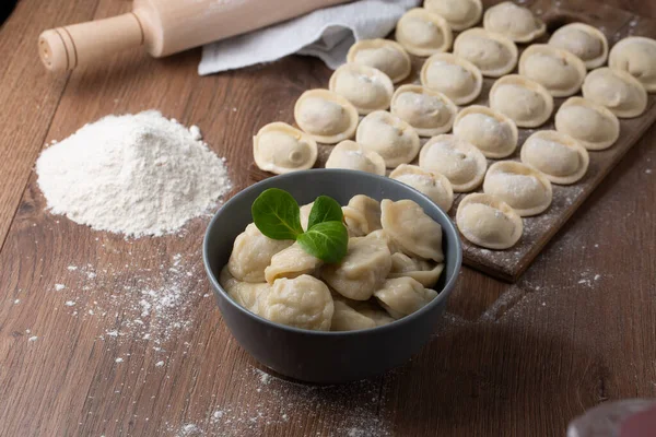 Zusammensetzung der grauen Schüssel mit zubereiteten russischen Fleischpelmeni mit Basilikumblatt und Holzbrett mit rohen Knödeln mit Hackfleisch auf dem Tisch mit Nudelholz, mit Mehl bestreut. Selektiver Fokus. — Stockfoto