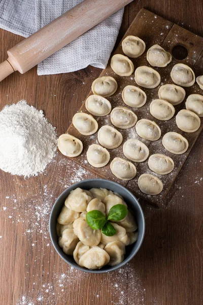 Composición de tazón gris con pelmeni de carne rusa preparada con hoja de albahaca y tablero de madera con albóndigas crudas con carne picada en la mesa con rodillo, espolvoreado con harina. Enfoque selectivo. —  Fotos de Stock