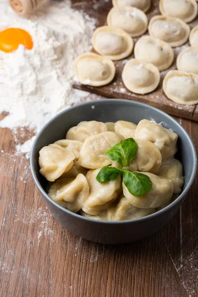 Composizione di ciotola grigia con pelmeni preparati di carne russa con foglia di basilico e bordo di legno con gnocchi crudi con carne macinata in tavola, cosparsa di farina. Focus selettivo. — Foto Stock