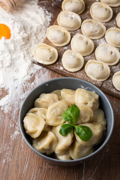 Composición de tazón gris con pelmeni de carne rusa preparada con hoja de albahaca y tablero de madera con albóndigas crudas con carne picada en la mesa, espolvoreada con harina. Enfoque selectivo. —  Fotos de Stock