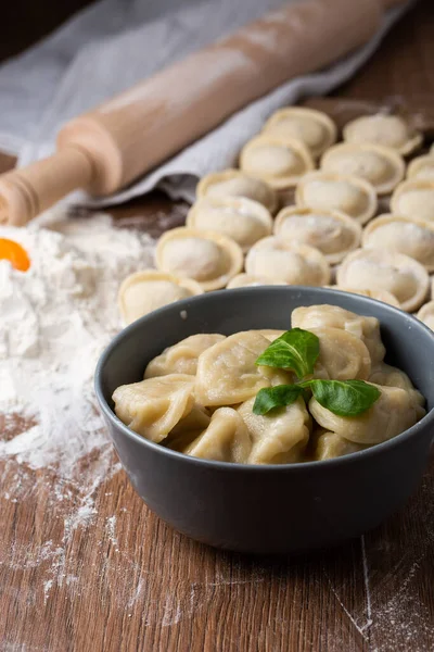 Composición de tazón gris con pelmeni de carne rusa preparada con hoja de albahaca y tablero de madera con albóndigas crudas con carne picada en la mesa, espolvoreada con harina. Enfoque selectivo. —  Fotos de Stock