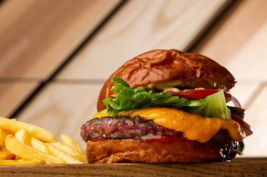 Tahta arka planda izole edilmiş patates kızartmalı büyük bir çizburger. Hamburger, köfteli soğan, domates, marul turşusu, erimiş çedar, hardal, domates reçeli, mayonez. Yatay