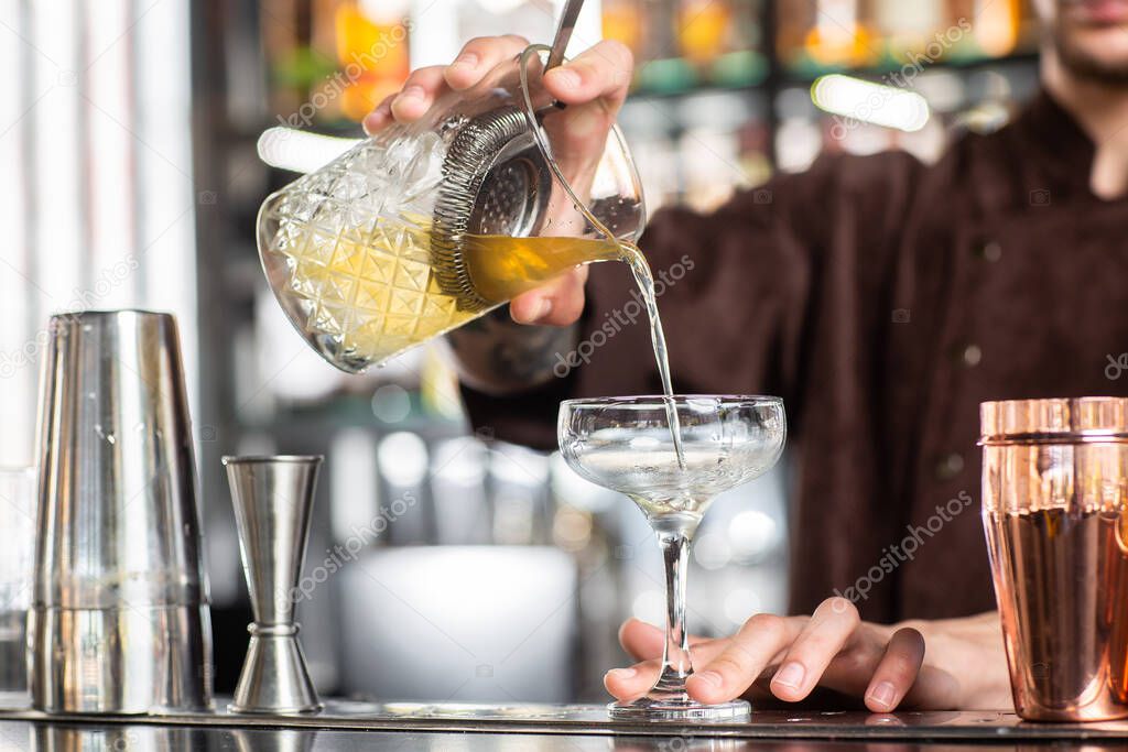 Bartender pouring an alcohol from the measuring glass cup through the strainer to the cocktail on the bar counter in the blurred background