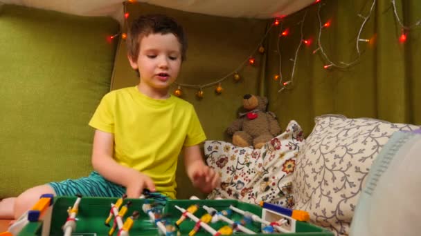 Υπέροχο Καυκάσιος Αγόρι Παίζοντας Ποδόσφαιρο τραπέζι σε ένα μικρό σπίτι με κουβέρτες και μαξιλάρια είναι διακοσμημένο με Χριστουγεννιάτικες γιρλάντες. Συναισθηματικό επιτραπέζιο παιχνίδι για την οικογένεια Pastime. Ψυχαγωγική δραστηριότητα στο σπίτι — Αρχείο Βίντεο