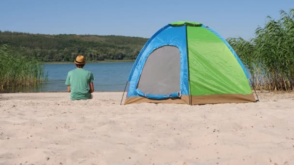 Retour gars vue reposant seul dans la tente de camping sur green eco travel. Vue arrière homme assis près de la tente touristique regardant la nature dans la journée calme d'été sur le lac de rive de sable ou rivière de rive après trekking ou randonnée locale — Video