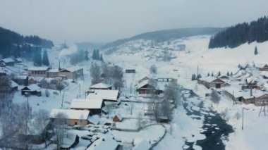 Donmuş bir nehrin kıyısındaki karlı bir köyün hava manzarası.