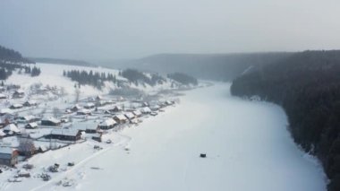 Donmuş bir nehrin kıyısındaki karlı bir köyün hava manzarası.